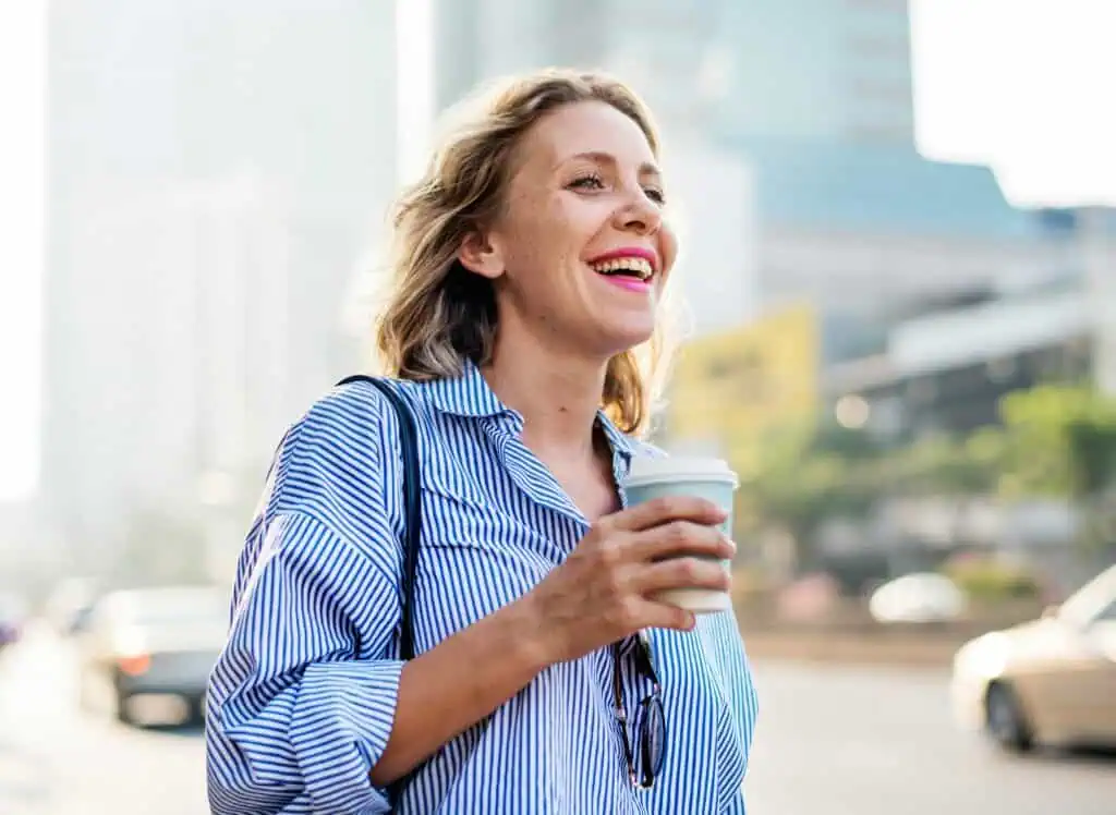 Lächelnde, selbstbewusste Frau in der Lebensmitte unterwegs in der Stadt mit Kaffeebecher – Symbol für Finanzplanung, Vermögensaufbau und Pensionsvorsorge für Frauen.